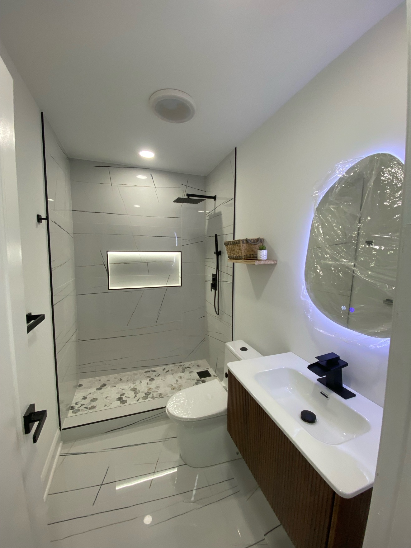 Bathroom with a marble-vein tile walk-in shower with a lit niche, a dark wood vanity and a backlit oval mirror