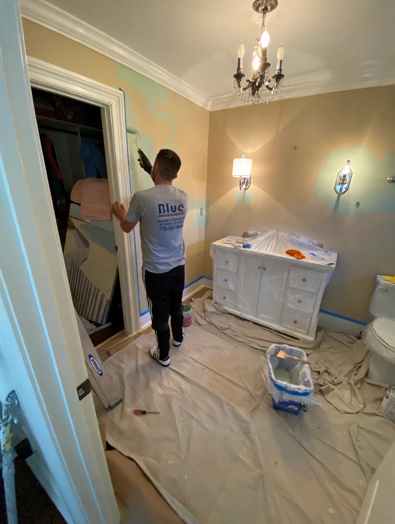 Blue Group painter in a branded shirt rolling a fresh coat in a bathroom — floors covered and the vanity protected with plastic